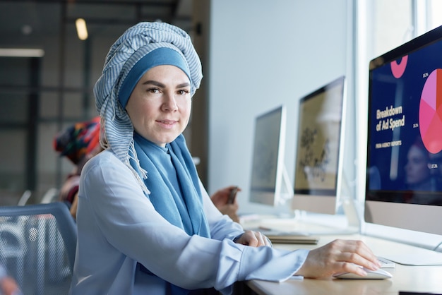 Portrait de femme en hijab assise sur son lieu de travail devant un écran d'ordinateur et regardant la caméra au bureau