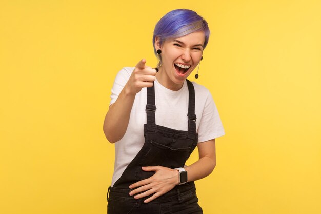 Portrait d'une femme hipster joyeuse et amusée avec des cheveux courts violets dans une salopette en jean tenant la main sur le ventre et riant bruyamment, pointant vers la caméra, vous narguant. isolé sur fond jaune, tourné en studio