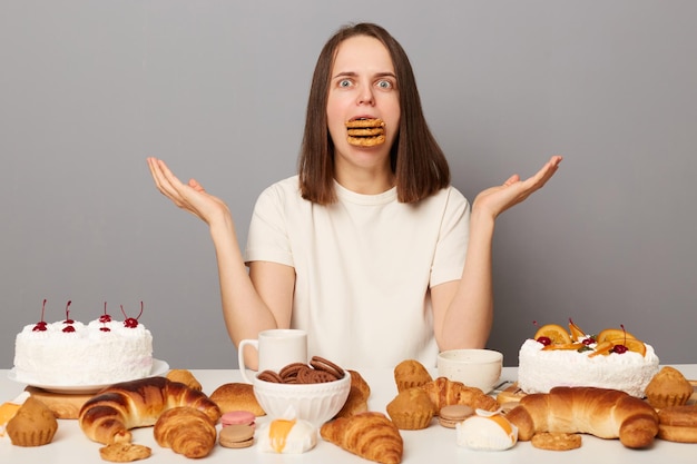 Portrait d'une femme incertaine confuse et choquée aux cheveux bruns portant un t-shirt blanc assis à table isolée sur fond gris haussant les épaules mangeant trop de bonbons a la bouche pleine de biscuits