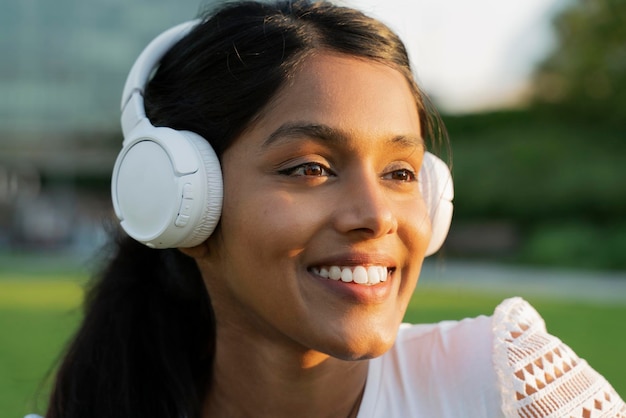 Portrait d'une femme indienne heureuse portant des écouteurs modernes blancs écoutant de la musique dans le parc