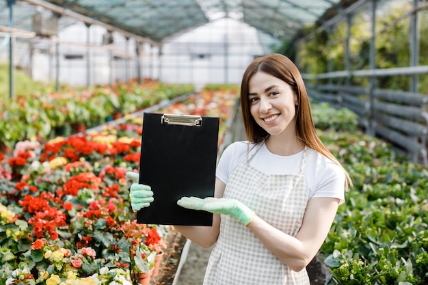 Portrait d'une femme jardinière au travail dans une serre avec un cahier examinant les fleurs en croissance dans la serre