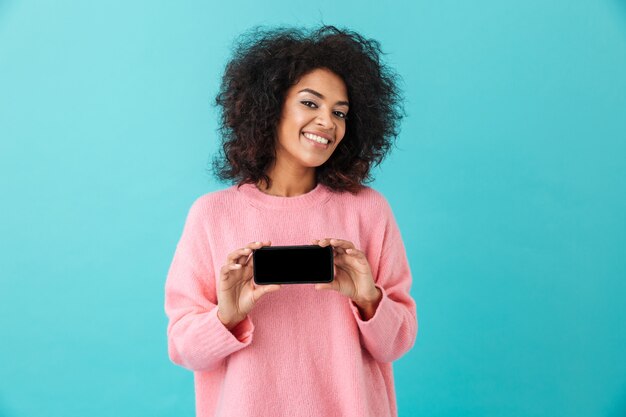 Portrait de femme joyeuse des années 20 tenant un téléphone portable et démontrant un écran noir copyspace, isolé sur mur bleu