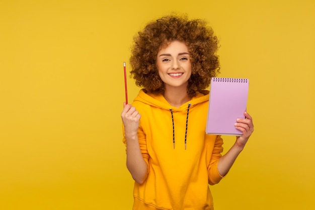 Portrait d'une femme joyeuse aux cheveux bouclés dans un sweat à capuche de style urbain à la recherche d'inspiration par une idée intelligente levant un crayon et un cahier pour écrire des plans liste de tâches prise de vue en studio à l'intérieur isolée sur fond jaune