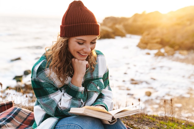 Portrait de femme joyeuse caucasienne lisant un livre et souriant alors qu'il était assis sur une couverture au bord de la mer