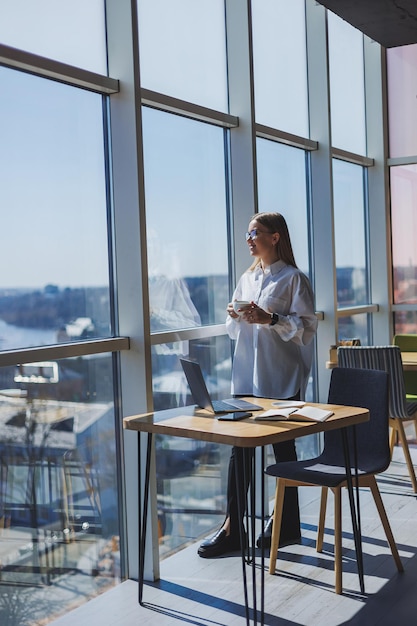 Portrait d'une femme joyeuse dans des verres classiques souriant pendant son temps libre dans un café avec une femme juive positive de café dans une chemise blanche bureau avec ordinateur portable travail à distance