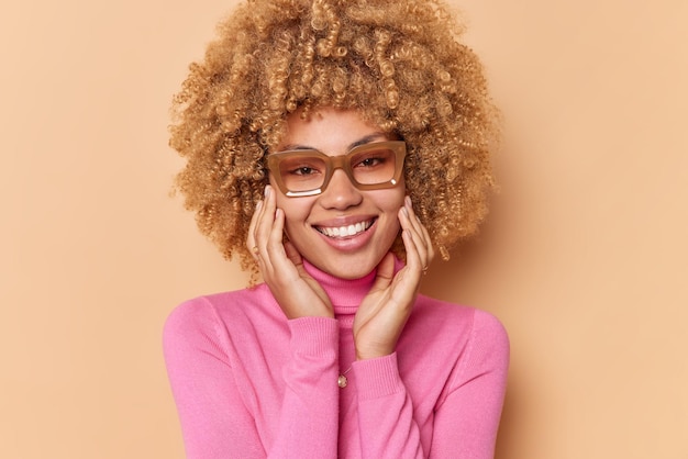 Portrait d'une femme joyeuse et heureuse touche le visage sourit doucement regarde directement la caméra se sent bien porte des lunettes et un col roulé isolé sur un mur de studio beige heureux d'entendre de bonnes nouvelles