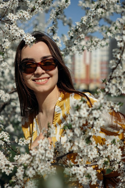 Portrait d'une femme joyeuse en lunettes de soleil et chemisier regardant la caméra dans le jardin de printemps