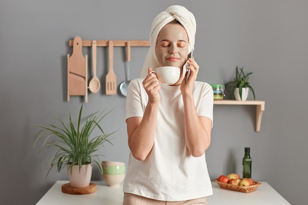 Portrait d'une femme joyeuse et satisfaite dans un masque cosmétique et avec une serviette sur la tête dans la cuisine sentant l'arôme agréable du café en dégustant une boisson chaude les yeux fermés et en parlant au téléphone intelligent