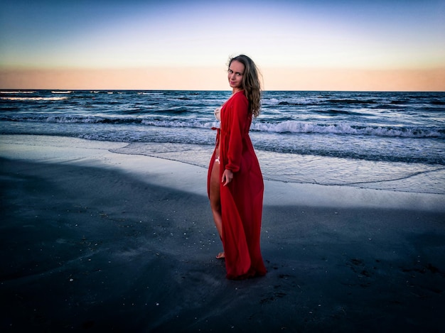 Photo portrait d'une femme en lingerie rouge marchant sur la plage contre le ciel au coucher du soleil