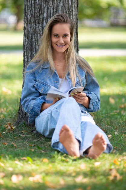 Portrait d'une femme lisant un livre assis dans le parc sur l'herbe, elle sourit et regarde vers le ca...