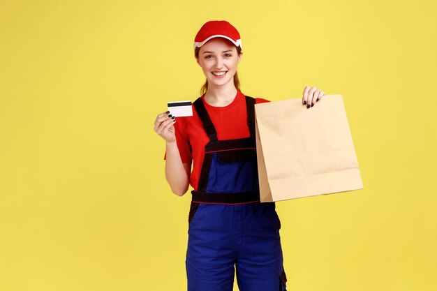 Portrait de femme de livraison tenant un colis papier et une banque de crédit regardant la caméra avec une expression heureuse portant une salopette et une casquette rouge Prise de vue en studio intérieure isolée sur fond jaune