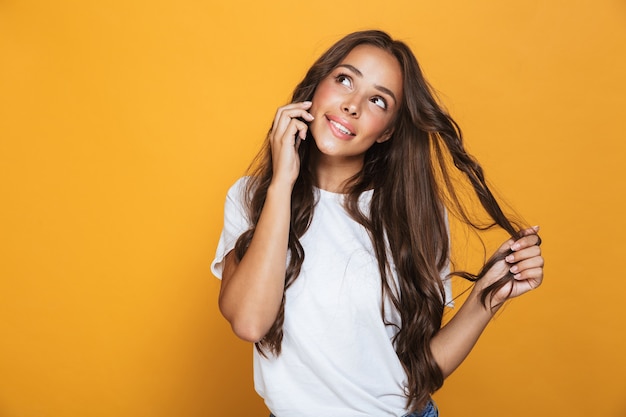 Portrait de femme magnifique des années 20 aux cheveux longs souriant et parlant au téléphone portable, isolé sur mur jaune