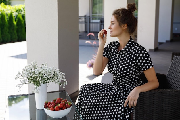 Portrait d'une femme magnifique portant une belle robe avec un motif à pois assis dans un patio et mangeant des fraises
