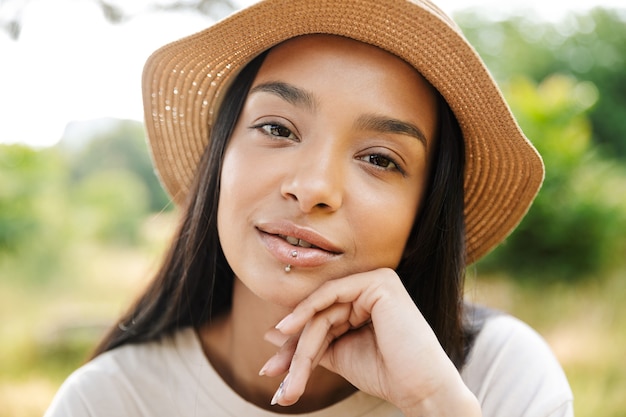 Portrait d'une femme magnifique portant un chapeau de paille et un piercing à la lèvre regardant la caméra en marchant dans un parc verdoyant