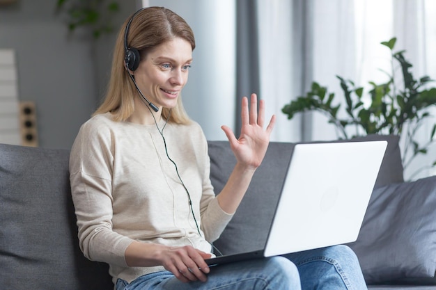 Portrait d'une femme à la maison assise sur le canapé et regardant la caméra et souriant accueille avec sa main utilise un casque et un ordinateur portable pour la communication vidéo