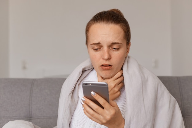 Portrait d'une femme malade avec un chignon assise sur un canapé enveloppé dans une couverture posant avec un téléphone portable dans les mains, souffrant de maux de gorge, composant le numéro du médecin, symptômes de la grippe.