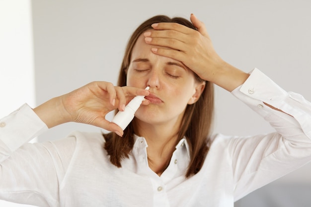 Portrait d'une femme malade et malsaine portant une chemise blanche de style décontracté, utilisant un spray nasal, gardant la main sur son front, debout les yeux fermés, ayant une température élevée.