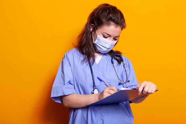 Portrait de femme avec masque facial et prenant des notes sur les fichiers du presse-papiers en studio. Assistant médical écrivant des informations avec un stylo sur des documents de manuels, ayant une protection contre le covid 19