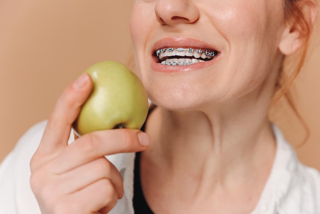 Portrait d'une femme mature avec des appareils orthopédiques sur les dents mangeant une pomme verte sur un fond beige