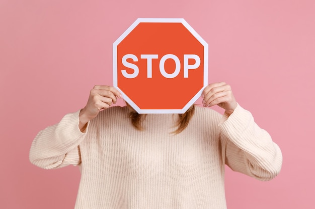 Portrait d'une femme méconnaissable cachant son visage avec un panneau rouge, campagne contre la violence domestique, protection des droits des femmes, portant un pull blanc. Tourné en studio intérieur isolé sur fond rose.