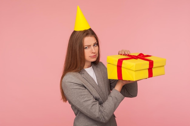 Portrait d'une femme mécontente bouleversée en costume d'affaires et avec un cône de fête sur la tête unboxing présent et regardant avec une drôle de grimace déçue insatisfaite du bonus cadeau tourné en studio intérieur isolé