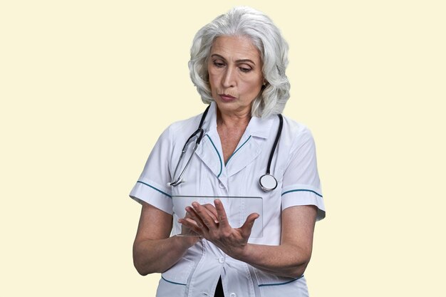 Portrait d'une femme médecin âgée sérieuse avec une maquette d'appareil transparent tablet pc imitation jaune pâle