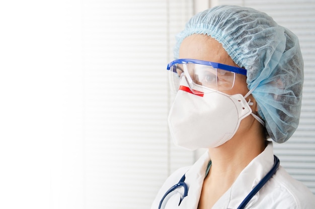 Photo portrait d'une femme médecin caucasienne dans des lunettes de masque et un chapeau avec sthétoscope sur fond de fenêtre blanche