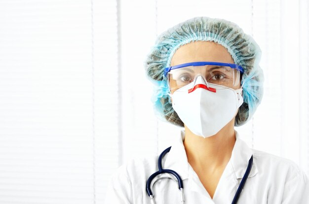 Photo portrait d'une femme médecin caucasienne dans des lunettes de masque et un chapeau avec sthétoscope sur fond de fenêtre blanche