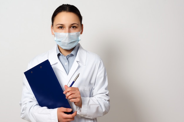 Portrait d'une femme médecin dans un masque de protection avec un presse-papiers et un stylo dans ses mains, établissant un dossier.