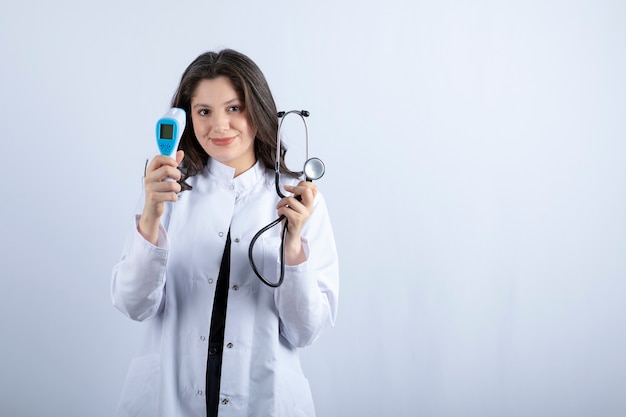 Portrait de femme médecin montrant un thermomètre et un stéthoscope sur un mur blanc.