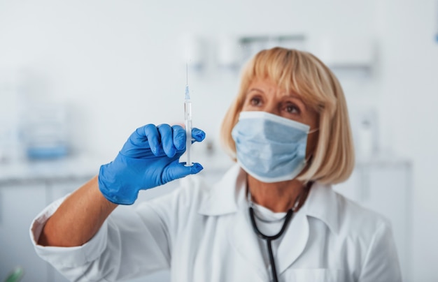 Portrait de femme médecin senior en masque avec seringue pleine de liquide.