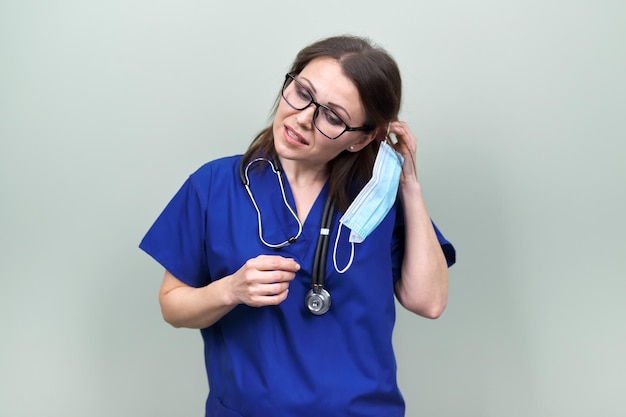 Portrait d'une femme médecin souriante et confiante avec stéthoscope en uniforme bleu. Femme enlevant le masque de protection médicale du visage, sur fond pastel vert