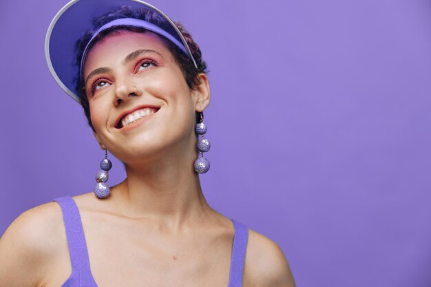 Portrait d'une femme de mode sportive posant souriante dans un costume de sport violet pour le yoga et une casquette transparente sur un fond violet monochrome Photo de haute qualité