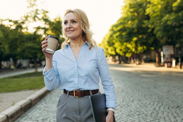 Portrait d'une femme mûre dans des vêtements élégants Femmes des années 50 Femme tenant un ordinateur portable et buvant du café tout en se promenant dans la ville