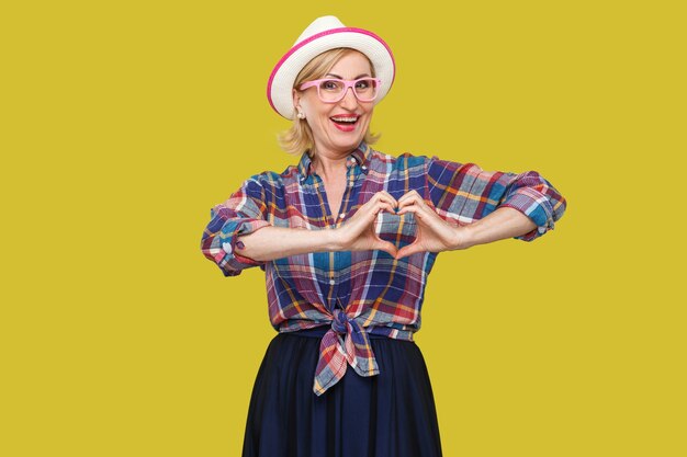 Portrait d'une femme mûre élégante et moderne paisible dans un style décontracté avec un chapeau blanc debout avec un geste en forme de coeur à la main, à la recherche et un sourire à pleines dents. tourné en studio intérieur isolé sur fond jaune