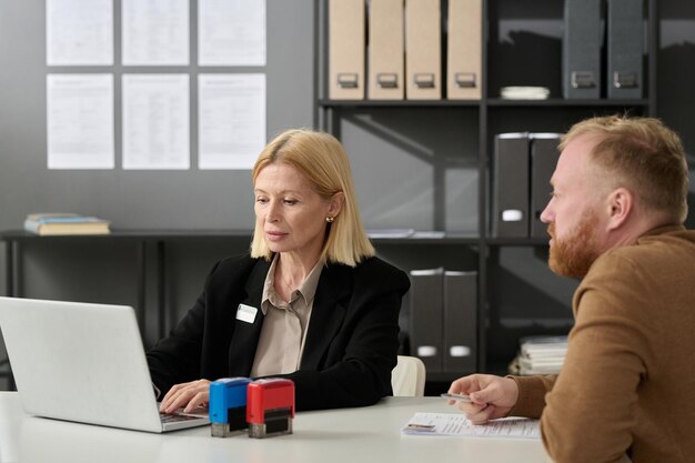 Portrait d'une femme mûre travaillant dans un bureau de services et utilisant un ordinateur portable tout en consultant un client masculin