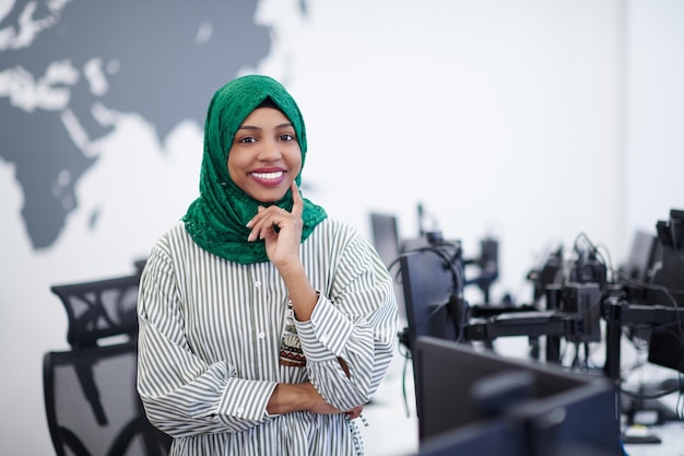 Portrait d'une femme musulmane africaine développeur de logiciels avec hijab vert debout au bureau de démarrage moderne à plan ouvert