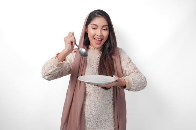 Portrait d'une femme musulmane asiatique affamée tenant des couverts et une assiette avec un espace de copie se sentant excitée à manger après le jeûne sur un fond blanc isolé concept du Ramadan