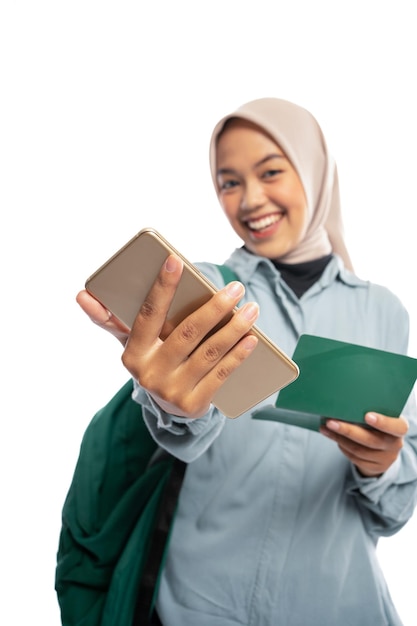 Photo portrait d'une femme musulmane asiatique souriante montrant un smartphone et un carnet de passe sur un fond blanc