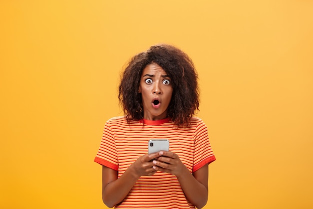 Portrait de femme à la peau foncée choquée avec une coiffure afro tenant un smartphone.