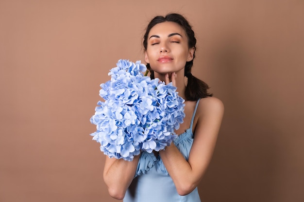 Portrait d'une femme à la peau parfaite et au maquillage naturel sur fond beige avec des nattes dans une robe tenant un bouquet de fleurs bleues