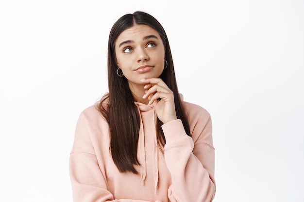 Portrait d'une femme pensive confuse, regardant le coin supérieur droit pensif, touchant le menton et faisant la moue perplexe, debout dans un sweat à capuche contre un mur blanc