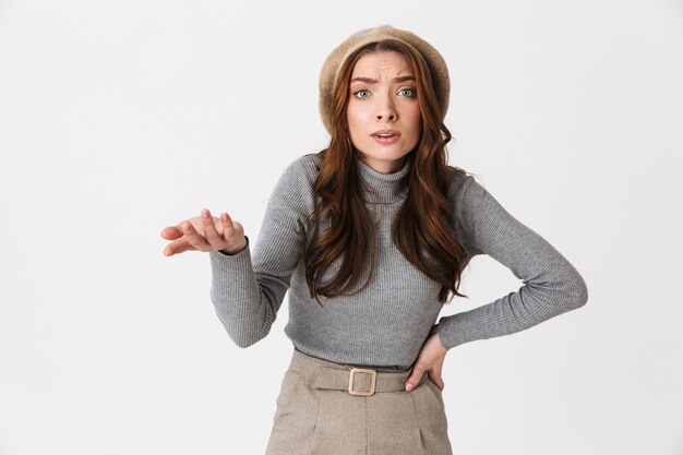 Portrait d'une femme perplexe portant un chapeau faisant des gestes et posant des questions isolées sur un mur blanc