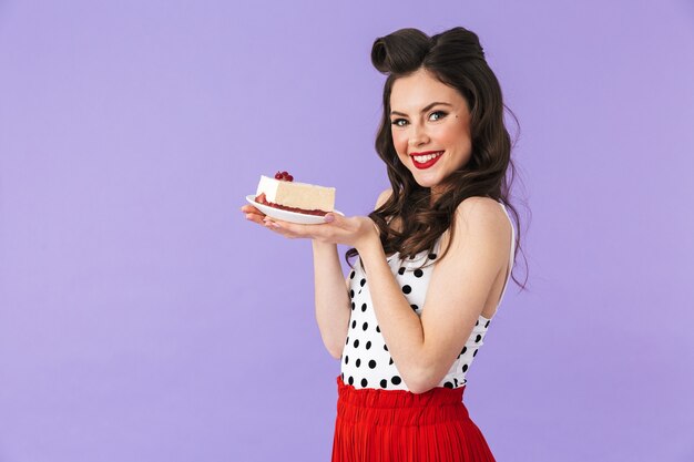 Portrait de femme pin-up positive en robe à pois vintage souriant tout en tenant et en mangeant un gâteau au fromage sucré isolé sur un mur violet