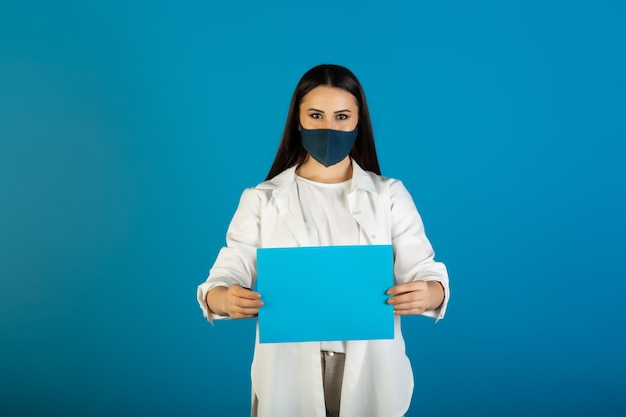 Portrait de femme portant un masque facial montre une feuille de papier vide bleue pour le texte. Mesure préventive contre le coronavirus COVID-19.