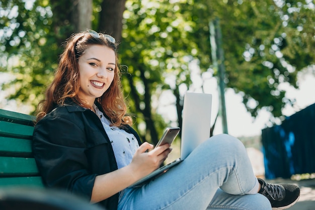 Portrait d'une femme positive de beau jeune corps regardant directement souriant alors qu'il était assis sur un banc avec un ordinateur portable sur les jambes et un smartphone à la main.