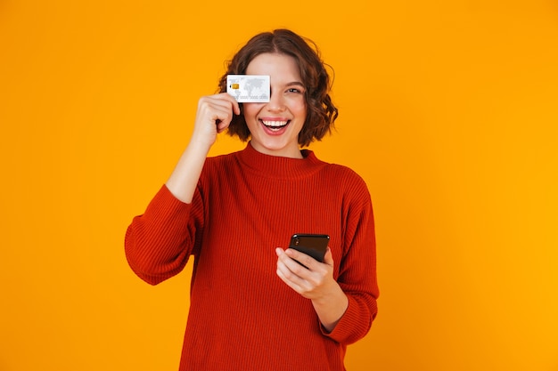 Portrait de femme positive portant chandail à l'aide de téléphone portable et carte de crédit en position debout isolé sur jaune