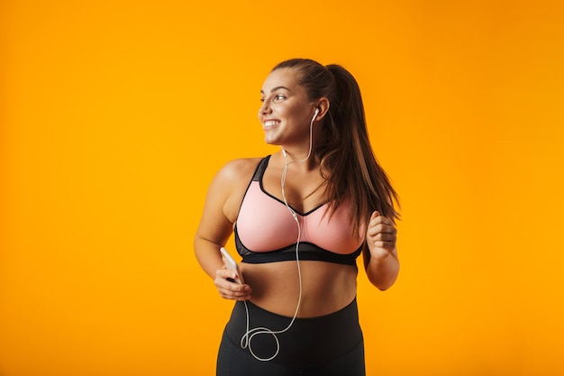 Portrait de femme potelée sportive en soutien-gorge sportif, écouter de la musique avec des écouteurs et smartphone, isolé sur fond jaune