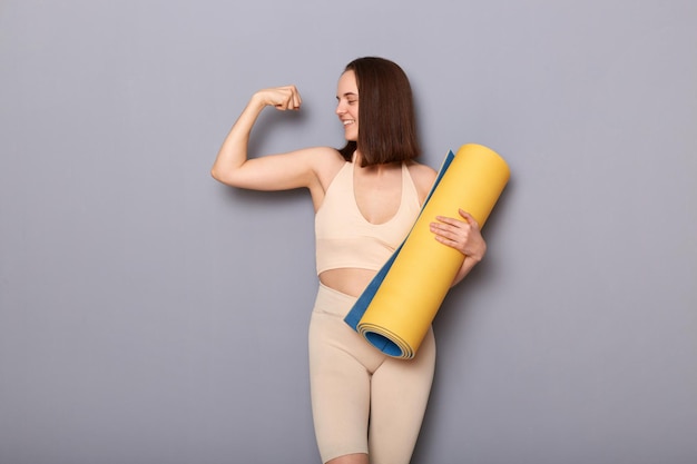 Portrait d'une femme prod confiante portant un haut beige et des leggins tenant un tapis de fitness isolé sur fond gris leva le bras montrant ses résultats de biceps après l'entraînement