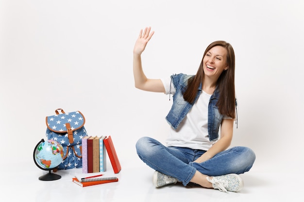 Portrait d'une femme qui rit heureuse dans des vêtements en denim étudiant agitant la main pour saluer assis près du globe, livres d'école de sac à dos isolés sur mur blanc